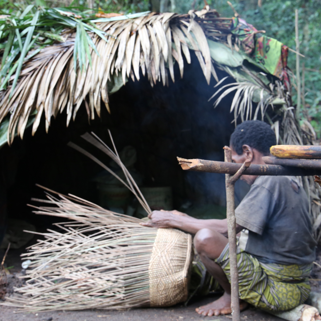 Man weaving basket beside a hut. 