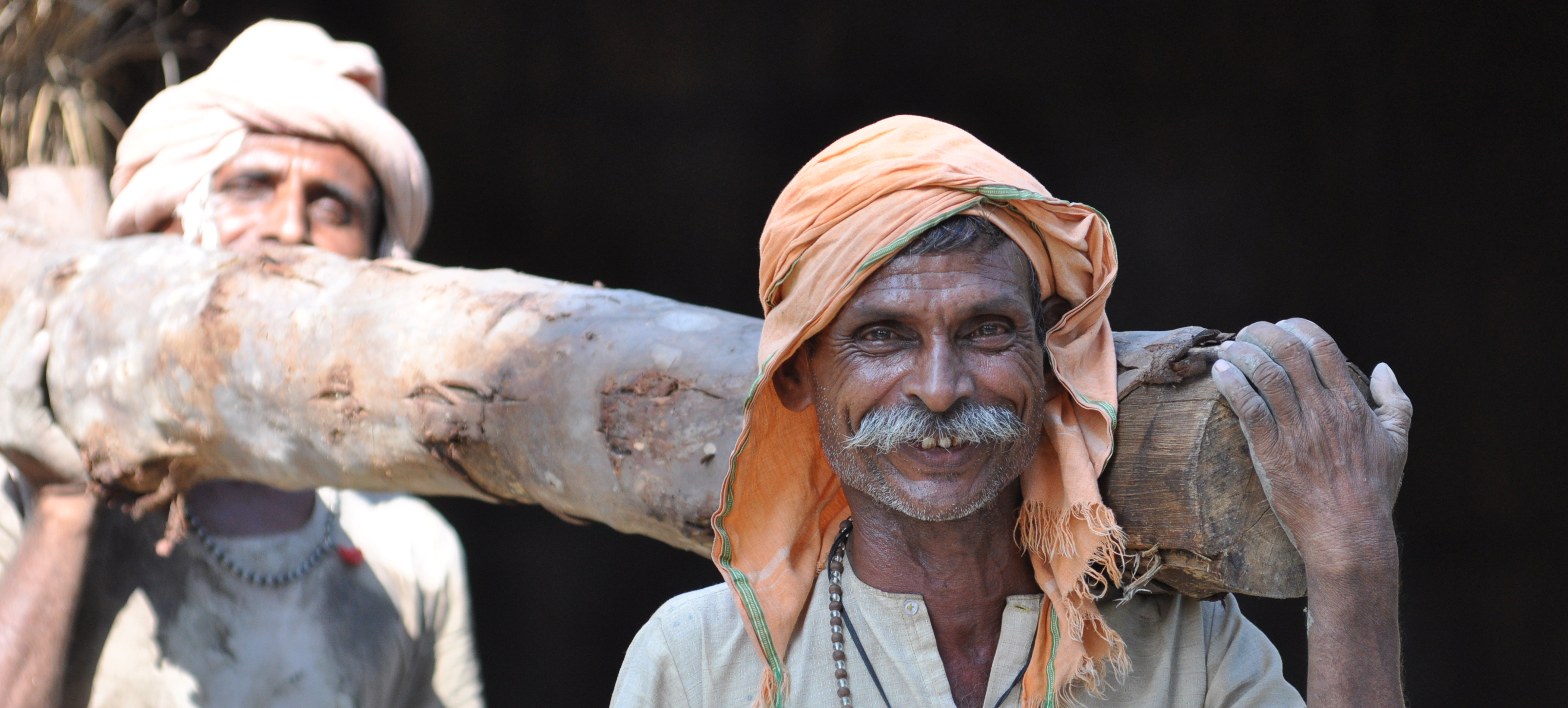two Indian men carry a felled log and smile for photo