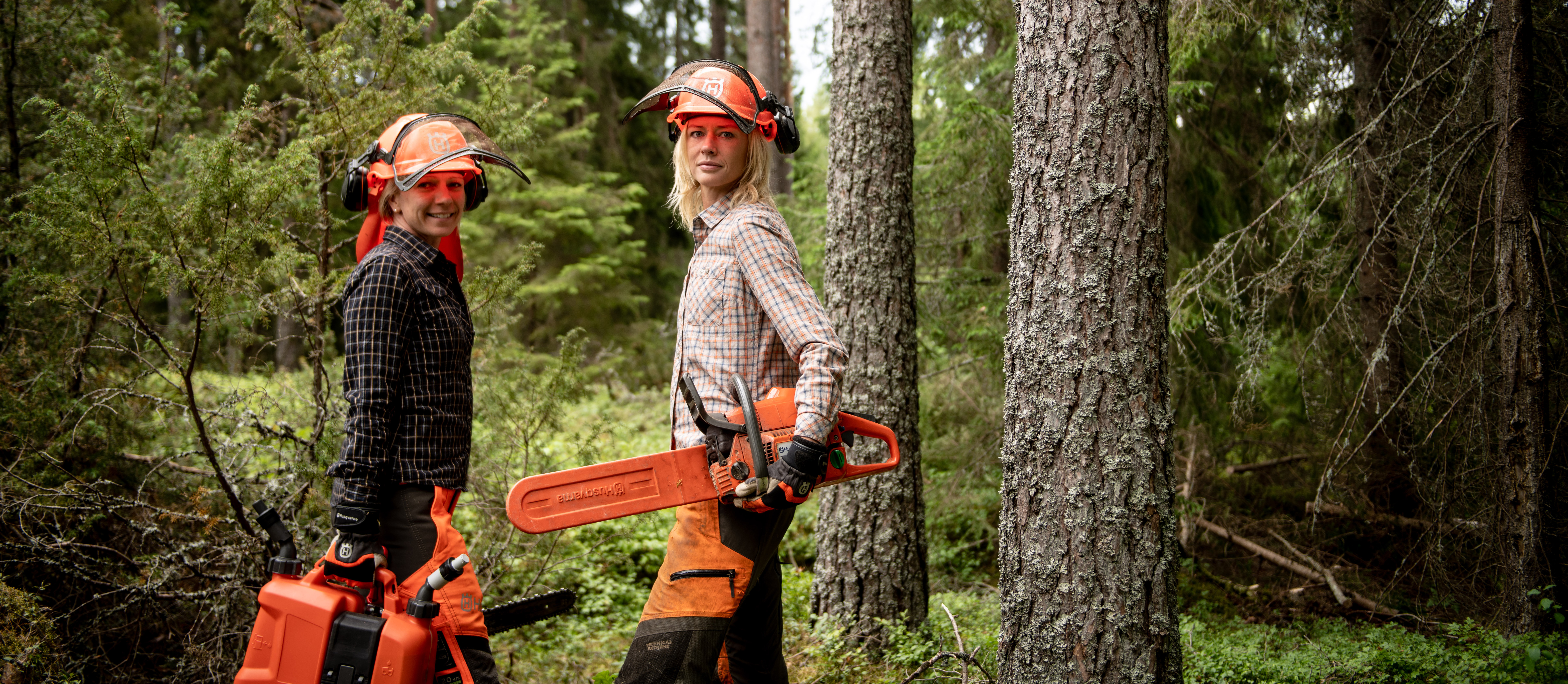 Two women standing in a forest carrying orange chainsaws