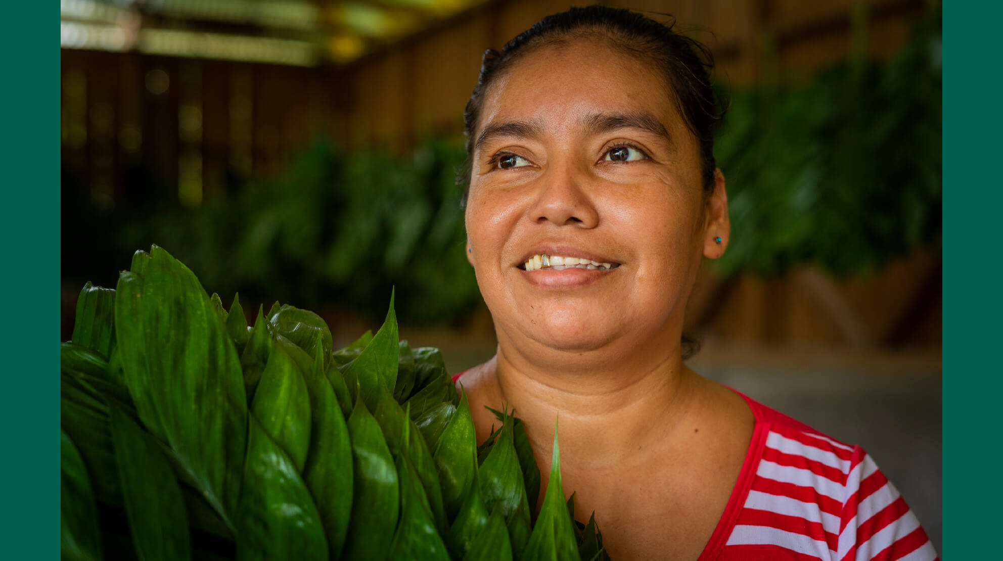 A smiling Indigenous woman holds a stack of palm leaves while looking in the distance.