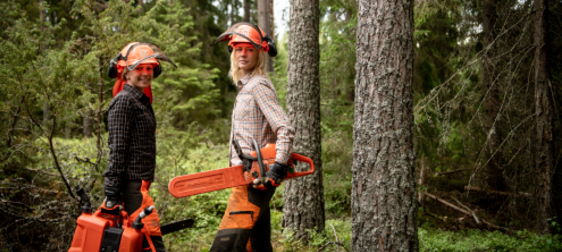 Two women standing in a forest carrying orange chainsaws