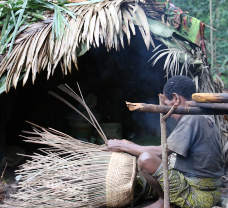 Man weaving basket beside a hut. 