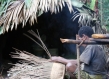 Man weaving basket beside a hut. 