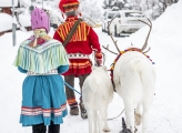 A man and woman in Sami traditional clothing walk in snow alongside bridled reindeer